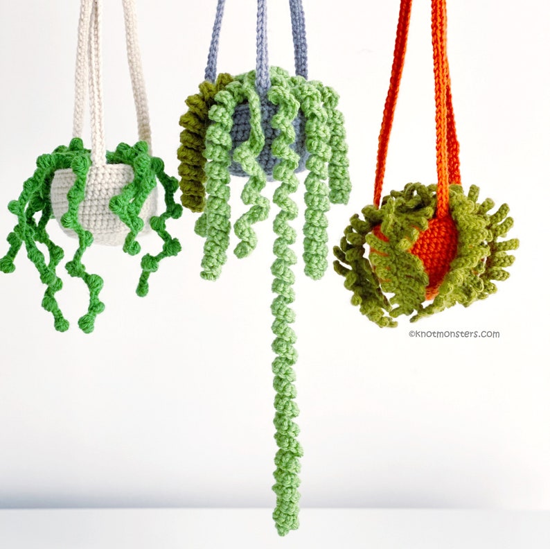 May include: Three crocheted hanging plant pots with green vines. The pots are white, light blue, and orange. The vines are made of green yarn and are hanging down from the pots.