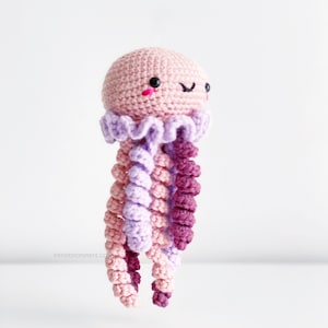 May include: A crocheted pink jellyfish with purple and pink tentacles. The jellyfish has a cute face with black eyes and a pink smile.