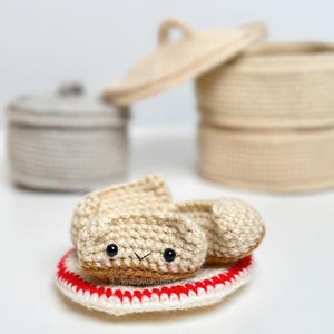 Könnte beinhalten: Ein gehäkelter Knödel mit einem lächelnden Gesicht, der auf einem weiß-rot gestreiften Untersetzer liegt. Der Knödel ist beige mit braunem Boden und schwarzen Augen. Im Hintergrund befinden sich zwei gehäkelte Körbe, einer mit Deckel.