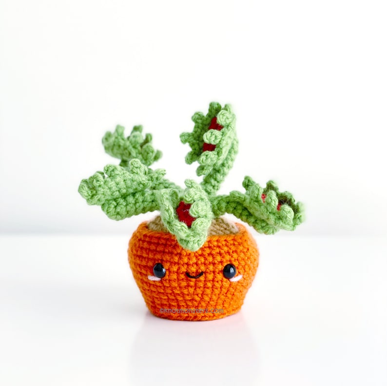 May include: A crocheted plush toy shaped like a Venus flytrap plant in a pot. The pot is orange and has a smiling face with black eyes. The plant has green leaves and red flowers.