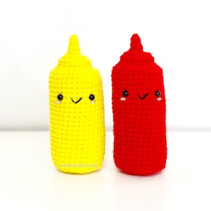 May include: Two crocheted plush toys shaped like condiment bottles. The bottle on the left is yellow and has a smiling face. The bottle on the right is red and also has a smiling face.