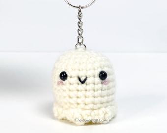May include: White crocheted ghost keychain with black eyes and pink cheeks. The ghost has a small smile and is attached to a silver key ring.