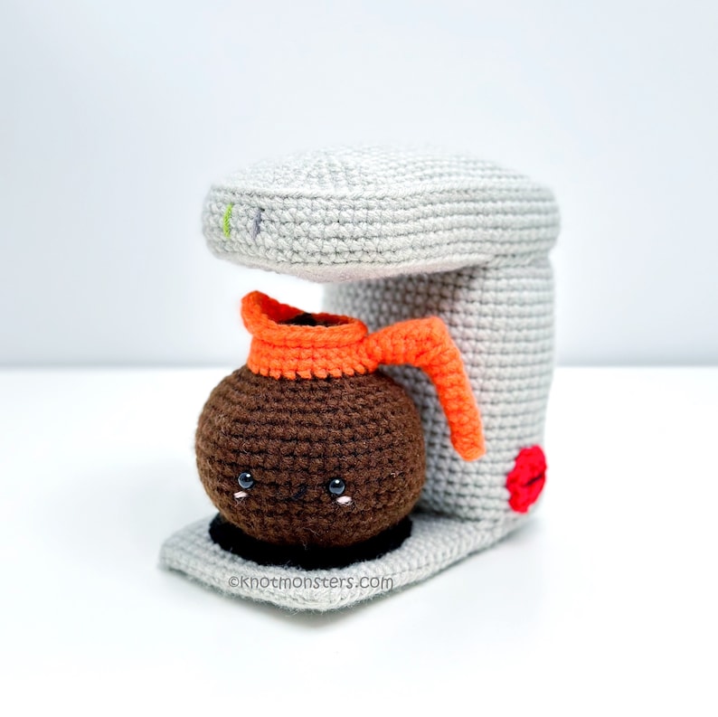 Pode incluir: Uma cafeteira de croch&ecirc; com uma cafeteira marrom com um rosto bonito e uma al&ccedil;a laranja. A cafeteira &eacute; cinza e tem um bot&atilde;o vermelho. O texto "knotmonsters.com" est&aacute; na parte inferior da cafeteira.