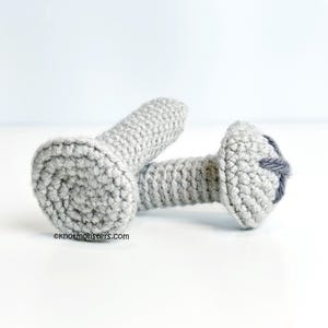 Pode incluir: Dois brinquedos de crochet em forma de parafuso, nas cores cinza e branco. Os brinquedos estão sobre uma superfície branca. O texto "knotmonsters.com" é visível no canto inferior esquerdo da imagem.