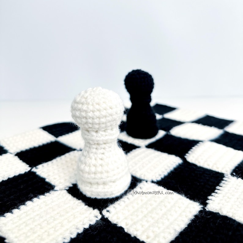 May include: A close-up of a crocheted chess set featuring a white pawn in the foreground and a black pawn in the background. The pieces are on a black and white checkered board. The website address "knotmonsters.com" is visible on the board.
