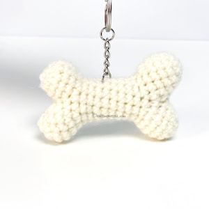 Può includere: Un portachiavi a forma di osso di cane in crochet bianco con una catena in metallo argentato e un anello in metallo argentato.