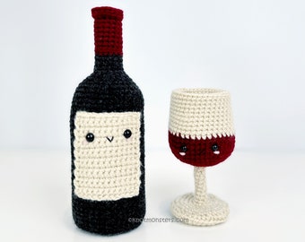 May include: A crocheted wine bottle and wine glass. The bottle is dark grey with a red top and a cream label. The wine glass is cream-coloured with a red wine fill. Both have black eyes and a smiling face.