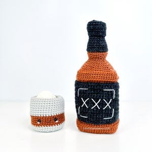 May include: A crocheted whiskey bottle and shot glass with a cute cartoon face. The bottle is brown and black with the text "XXX" on the label. The shot glass is gray with a brown rim and a brown liquid inside.