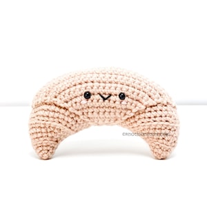 May include: A crocheted croissant plush toy with a smiling face. The croissant is light brown and has black eyes and a small mouth.