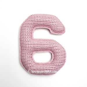 Puede incluir: Un número seis de ganchillo rosa claro hecho a mano, tumbado sobre un fondo blanco. El número es suave y afelpado, con una forma redondeada y una gran abertura en el centro. La dirección del sitio web "knotmonsters.com" es visible en la parte inferior.