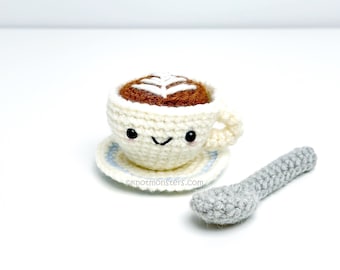 May include: A crocheted coffee cup with a smiling face, holding a brown coffee with a white foam design. The cup is on a white saucer with a blue trim. A gray crocheted spoon is next to the cup.