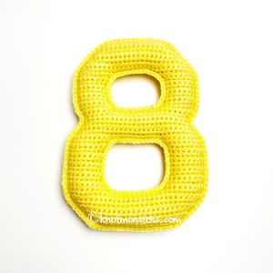 Puede incluir: Un número ocho amarillo brillante, tejido a ganchillo, con dos círculos abiertos. El número hecho a mano se muestra sobre un fondo blanco. El texto "©knotmonsters.com" es visible en la parte inferior de la imagen.