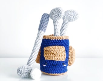 May include: A crocheted golf bag with a blue and brown exterior and a smiling face. The bag has three golf clubs sticking out of the top, all crocheted in a light gray color.