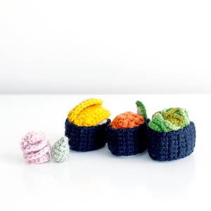 May include: A selection of handmade crochet sushi pieces. The sushi rolls are dark blue with various colourful toppings, including yellow, orange, and green. Two smaller, pink and green crochet pieces are also present. The items are arranged on a white surface.