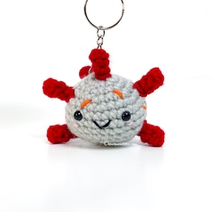 Puede incluir: Un llavero de axolotl de crochet gris con extremidades rojas y ojos negros. El axolotl sonríe y tiene cejas naranjas. El llavero está unido a un llavero plateado.