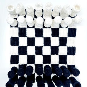 May include: A crocheted chess set with a black and white checkered board. The chess pieces are white, and the black pieces are attached to the bottom of the board. The website address "knotmonsters.com" is visible on the board.
