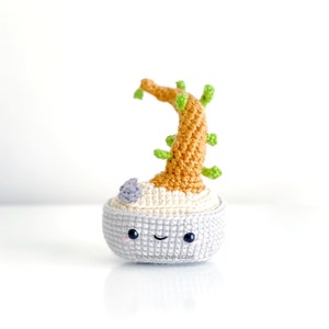 Peut inclure: Une plante crochetée avec un visage souriant. La plante a un tronc brun clair avec des feuilles vertes et est dans un pot gris. Une petite fleur violet clair est sur le côté. Le pot a un visage souriant avec des yeux noirs et une bouche incurvée. Le texte "knotmonsters.com" est sur le pot.