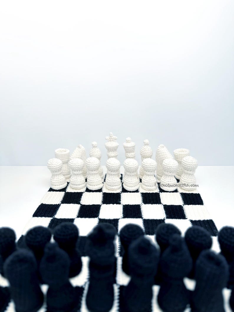May include: A crocheted chess set with black and white pieces arranged on a checkered board. The white pieces are in focus, while the black pieces are blurred in the foreground. The board is black and white.