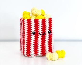 May include: A crocheted popcorn bucket with a red and white striped pattern and a smiling face. The bucket is filled with yellow crocheted popcorn pieces. There are also a few popcorn pieces on the white surface below the bucket.