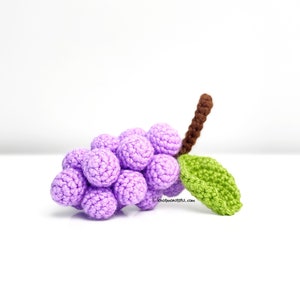 May include: A crocheted bunch of purple grapes with a brown stem and a green leaf. The grapes are made of yarn and are about the size of a golf ball. The stem is about 7.5 cm long. The leaf is about 5 cm wide. The grapes are arranged in a cluster. The stem is attached to the top of the cluster. The leaf is attached to the side of the cluster. The grapes are a light purple colour. The stem is a brown colour. The leaf is a green colour. The grapes are a soft, fuzzy texture. The stem is a smooth, hard texture. The leaf is a soft, smooth texture.