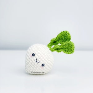 May include: A crocheted white radish with a smiling face and green leaves. The radish has small black eyes and pink cheeks. The website address "knotmonsters.com" is printed on the bottom left.