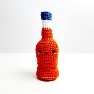 May include: A handmade, orange crochet bottle with a smiling face, a blue, white, and blue top, and the text "eknotmonsters.com" at the bottom. The bottle is a whimsical, handcrafted item.