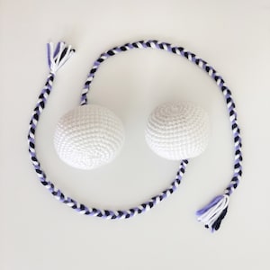 Może przedstawiać: Dwie białe kule wykonane na szydełku połączone długim, plecionym sznurkiem w paski fioletowe, białe i czarne. Sznurek ma po obu stronach frędzle.