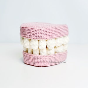 Puede incluir: Un conjunto de dientes de ganchillo rosa y blanco. Los dientes están hechos de hilo blanco y las encías de hilo rosa.
