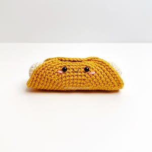 May include: A crocheted taco with a smiling face. The taco shell is mustard yellow, with white accents on the ends. The face has black eyes and pink cheeks. The taco is a handmade item.