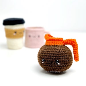 Pode incluir: Uma cafeteira de croch&ecirc; marrom com uma tampa e uma al&ccedil;a de croch&ecirc; laranja. A cafeteira tem um rosto bonito com olhos pretos e um pequeno sorriso. A cafeteira est&aacute; sentada em uma superf&iacute;cie branca.