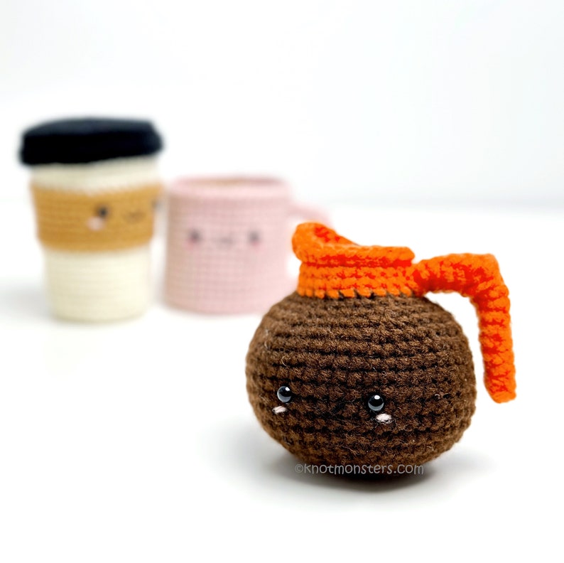Pode incluir: Uma cafeteira de croch&ecirc; marrom com uma tampa e uma al&ccedil;a de croch&ecirc; laranja. A cafeteira tem um rosto bonito com olhos pretos e um pequeno sorriso. A cafeteira est&aacute; sentada em uma superf&iacute;cie branca.