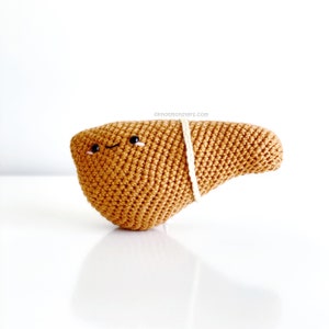 May include: A crocheted brown liver plush toy with a white string tied around it. The toy has a smiling face with black eyes and pink cheeks. The text "knotmonsters.com" is visible in the background.