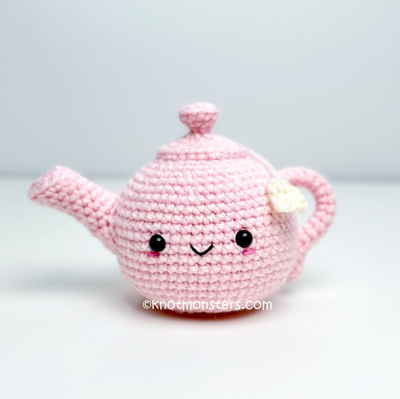 Pode incluir: Uma chaleira de croch&ecirc; rosa com um rosto sorridente e um saquinho de ch&aacute; branco. A chaleira tem um pequeno bico e uma al&ccedil;a.