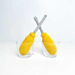 May include: Two crochet screwdrivers, each in a clear glass. The screwdrivers have yellow handles and grey tops. The screwdrivers are arranged in an X shape. The website address knotmonsters.com is visible on the bottom of the glass.