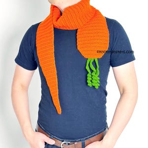 May include: An orange crochet scarf shaped like a carrot, with green leafy ends. The scarf is worn around the neck and hangs down the front. The scarf is made of a soft, textured yarn.