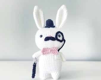 May include: A white crocheted bunny wearing a black top hat, a black mustache, a pink bow tie, and a black monocle. The bunny is holding a black and white walking stick.