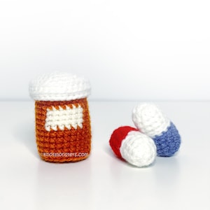 Puede incluir: Botella de pastillas naranja tejida con una etiqueta y tapa blancas, junto a dos pastillas tejidas, una roja y blanca, la otra azul y blanca. El sitio web knotmonsters.com es visible en la botella.