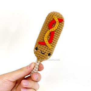 Peut inclure: Un corndog en crochet avec un visage souriant, de la moutarde jaune et du ketchup rouge. Le corndog est tenu dans une main.
