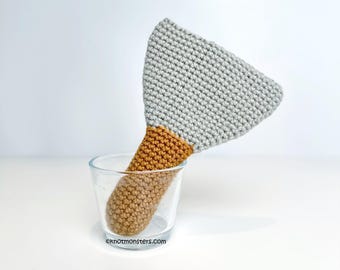 May include: A gray and brown crocheted spatula toy in a clear glass. The spatula is shaped like a triangle with a rounded handle.