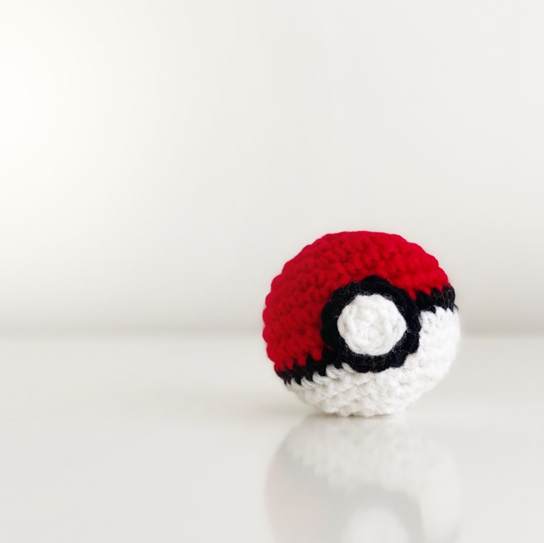 Puede incluir: Una bola de crochet roja y blanca con un c&iacute;rculo negro en el centro. La bola es una r&eacute;plica de una Pok&eacute; Ball.
