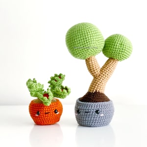 May include: Two crocheted potted plants with smiling faces. The larger plant has a brown stem and two green round leaves. The smaller plant is orange with green leaves. Both plants are in grey pots.
