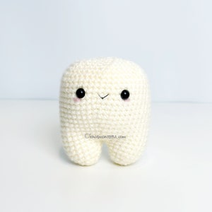 Peut inclure: Un jouet en peluche en forme de dent blanche en crochet avec des yeux noirs et une bouche souriante. Le jouet est debout sur une surface blanche.