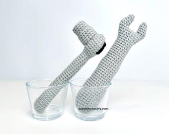 May include: Two gray crocheted toy tools, a wrench and a hammer, are standing upright in two clear glass cups. The cups are on a white surface.