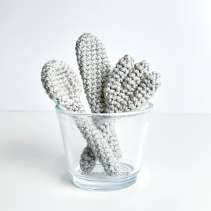 Puede incluir: Un vaso transparente con varias formas de cactus de ganchillo grises en su interior. Los cactus tienen un aspecto texturizado y están dispuestos para parecerse a una pequeña escena desértica. El fondo es blanco liso.
