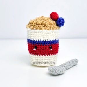 Puede incluir: Un postre de ganchillo con una cara sonriente, con una parte superior beige, una base blanca y rayas rojas y azules. Está rematado con bayas de ganchillo rojas y azules. Una cuchara de ganchillo gris descansa a su lado. La dirección del sitio web "knotmonsters.com" es visible.