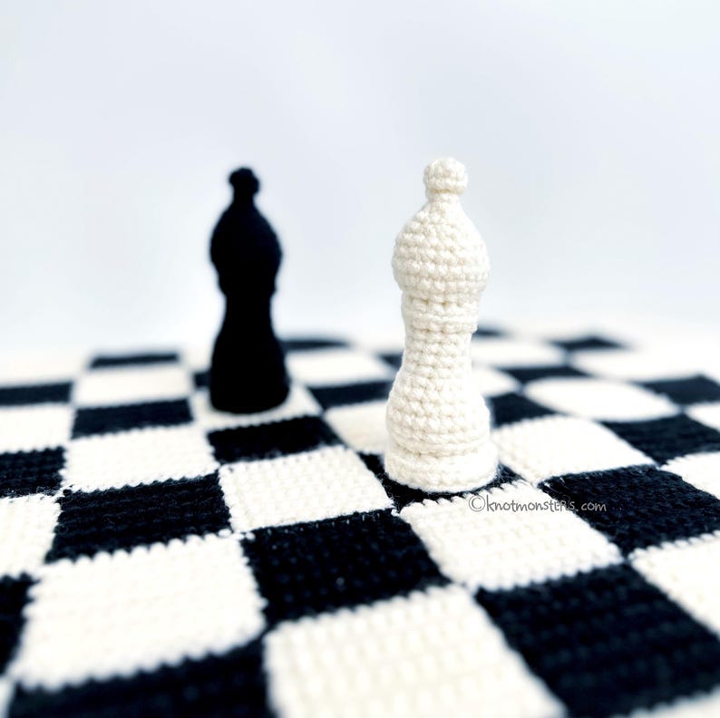 May include: A crocheted chess set featuring a black bishop and a white bishop on a black and white checkered board. The pieces are handmade and have a textured appearance. The background is white, highlighting the chess pieces.