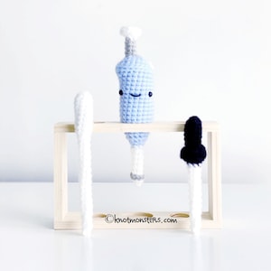 Puede incluir: Una pipeta de crochet azul con una cara sonriente, una pipeta de crochet blanco y una pipeta de crochet negro se muestran en un soporte de tubos de ensayo de madera. El texto "knotmonsters.com" es visible en la esquina inferior derecha de la imagen.