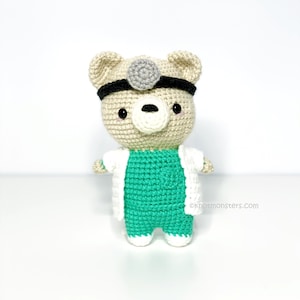 Puede incluir: Un oso de peluche tejido a crochet vestido de médico, con una bata blanca, un top y pantalones de cirujano verdes, y una diadema con una linterna frontal. El oso tiene una nariz blanca y ojos negros.