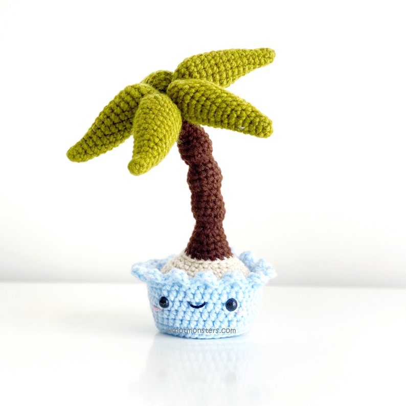 May include: A crocheted palm tree toy with a brown trunk and green leaves. The tree is planted in a blue pot with a smiling face.