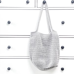 May include: A gray crocheted tote bag hanging on a white dresser with black knobs. The bag has a wide, flat bottom and two long straps.